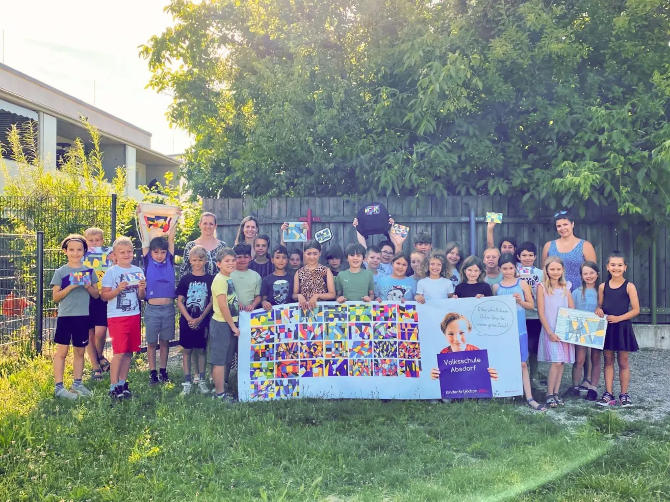 Eine Gruppe von Kindern und Erwachsenen steht in einem Garten mit einem bunten Banner vor ihnen. Sie halten verschiedene Gegenstände und scheinen für ein Foto zu posieren. Das Banner hat ein farbenfrohes Design und die Worte 'Volksschule Absdorf' darauf. Hinter ihnen steht ein Holzzaun mit einem Gebäude.