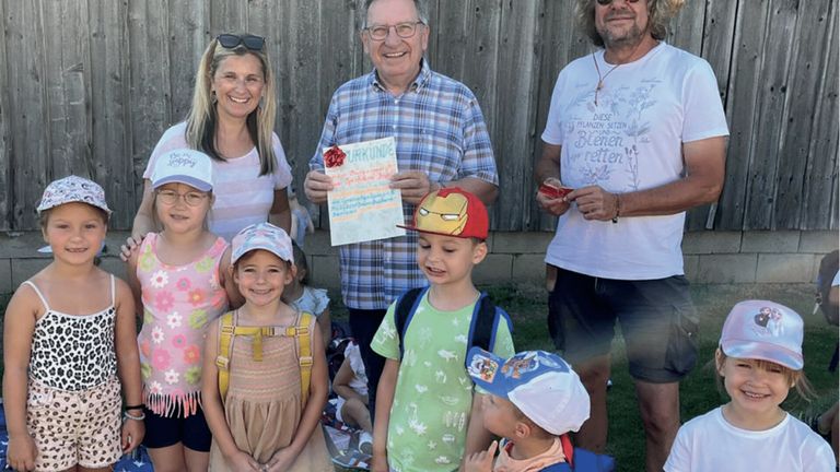 Eine Gruppe lächelnder Erwachsener und Kinder steht draußen. Ein Mann hält eine Karte mit dem Text 'Urkunde'. Ein junger Junge trägt einen Iron Man Hut. Sie scheinen für ein Foto zu posieren.