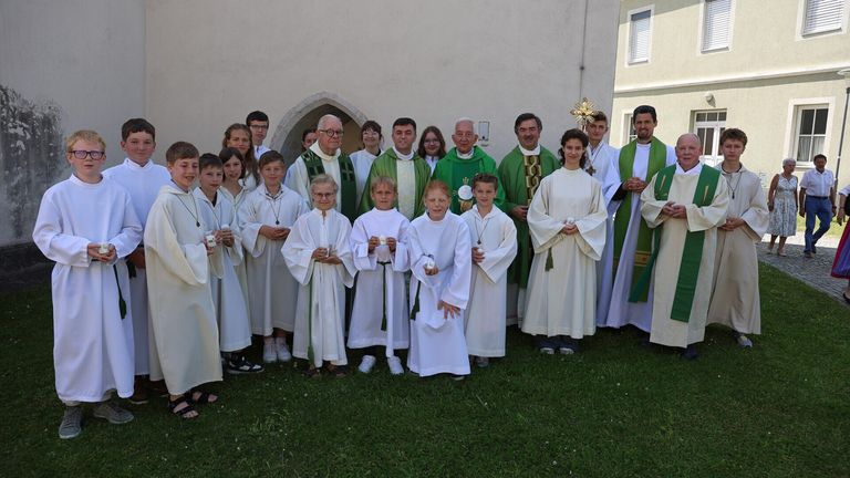 Eine Gruppe von Menschen, wahrscheinlich Priester und Messdiener, steht gemeinsam auf einem Rasen. Sie tragen weiße Gewänder und halten Kerzen. Im Hintergrund befindet sich ein Gebäude mit einem Bogengang.