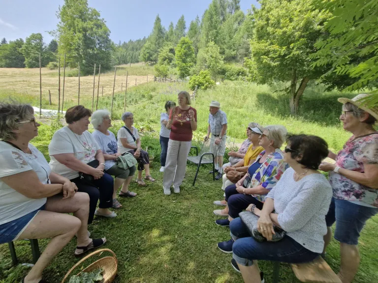 Eine Zusammenkunft älterer Frauen im Freien. Eine Frau steht in der Mitte und spricht. Die Frauen sitzen kreisförmig auf einem Rasen, mit Bäumen und Sträuchern im Hintergrund.