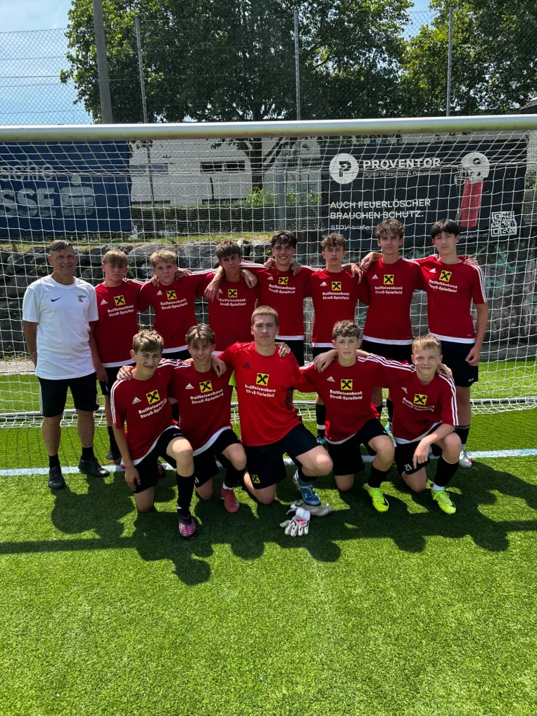 Eine Fußballmannschaft in roten Trikots mit weißen Akzenten posiert für ein Foto vor einem Tor. Der Trainer in einem weißen Shirt steht neben ihnen. Der Platz hat Kunstrasen.