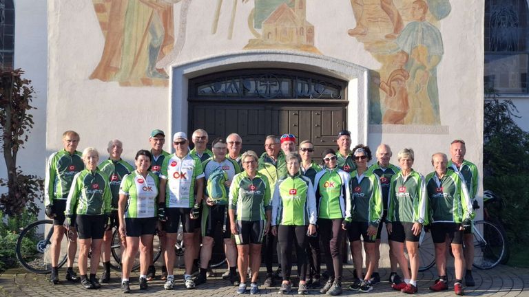 Eine Gruppe von Radfahrern, gekleidet in grünen und weißen Trikots mit verschiedenen Logos, steht vor einem Gebäude mit einer Holztür. Die Radfahrer posieren für ein Gruppenfoto.