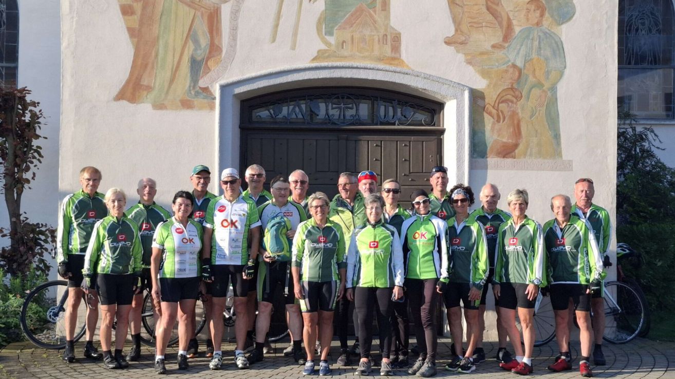 Eine Gruppe von Radfahrern, gekleidet in grünen und weißen Trikots mit verschiedenen Logos, steht vor einem Gebäude mit einer Holztür. Die Radfahrer posieren für ein Gruppenfoto.