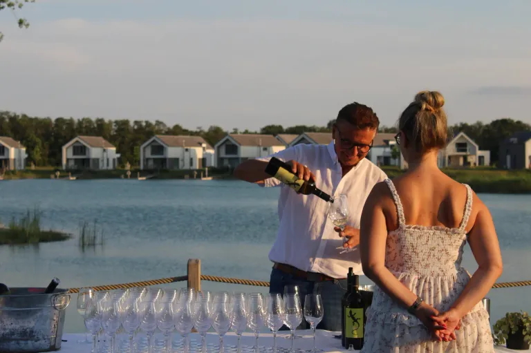 Ein Mann schenkt einer Frau Wein in ein Glas ein, am See mit Häusern im Hintergrund.