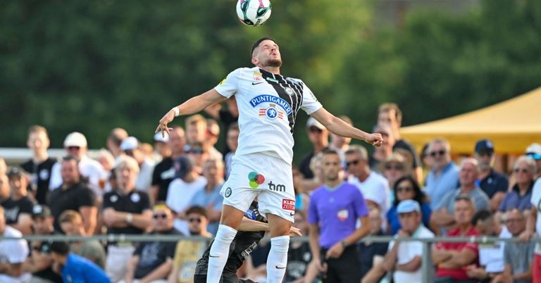 Ein Fußballspieler in einem weißen Trikot mit dem Wort Puntigamer springt hoch, um den Ball zu köpfen. Zuschauer im Hintergrund schauen dem Spiel zu.