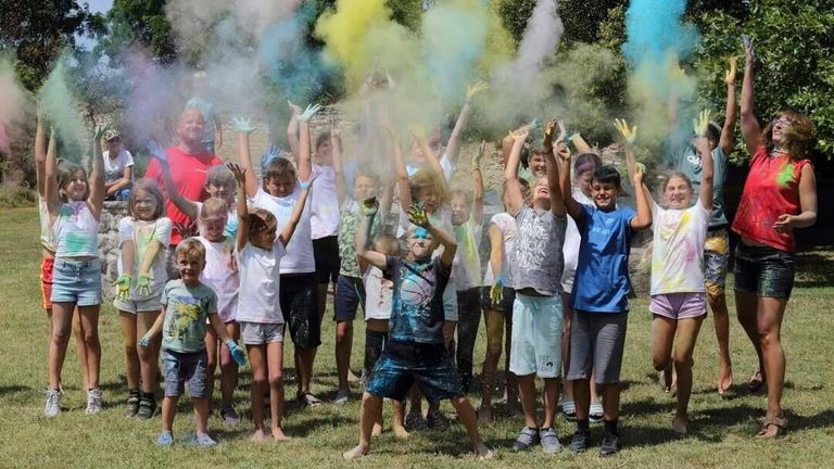 Eine Gruppe von Kindern veranstaltet draußen einen farbenfrohen Pulverkampf. Sie lächeln und heben die Hände in die Luft. Einige tragen Handschuhe. Bäume und Gras sind im Hintergrund.