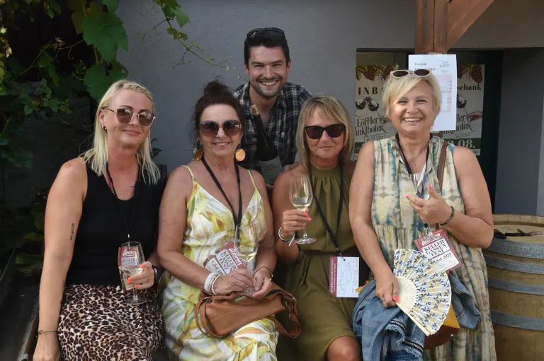 Fünf Erwachsene, vier Frauen und ein Mann, lächeln und posieren für ein Foto. Sie halten Weingläser und tragen Sonnenbrillen. Die Frau auf der rechten Seite hält einen Ventilator. Hinter ihnen ist eine Wand mit einer Weinrebe und ein Schild, das "Löffler Inkhlöfer" steht.