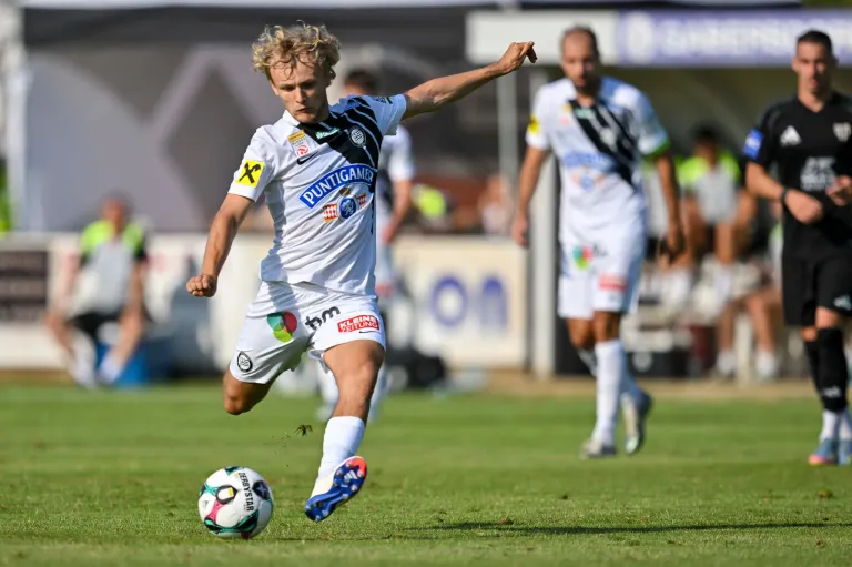 Ein Fußballspieler in weißer Kleidung mit 'PUNTIGAMER' auf der Vorderseite tritt den Ball, während ein anderer Spieler im Hintergrund folgt.
