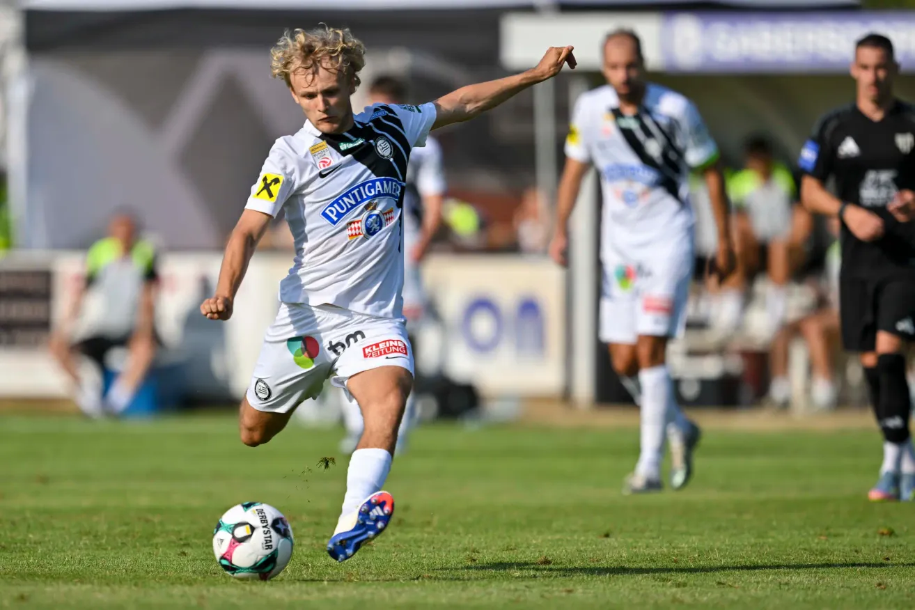 Ein Fußballspieler in weißer Kleidung mit 'PUNTIGAMER' auf der Vorderseite tritt den Ball, während ein anderer Spieler im Hintergrund folgt.