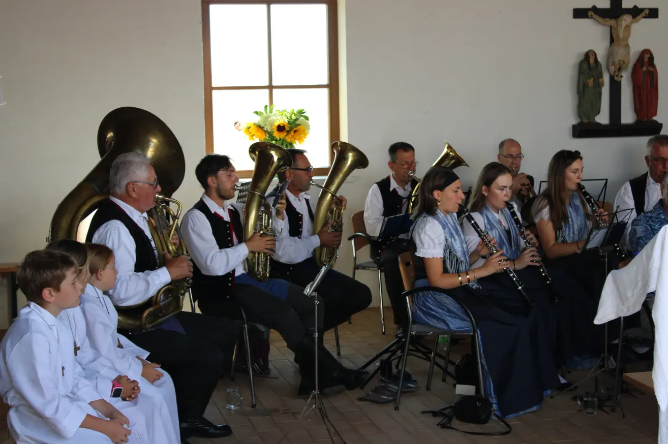 Eine Gruppe von Musikern spielt Instrumente in einem Raum mit gefliestem Boden. Zwei Frauen spielen sitzend die Klarinette. Männer spielen stehend Tubas und Posaunen.