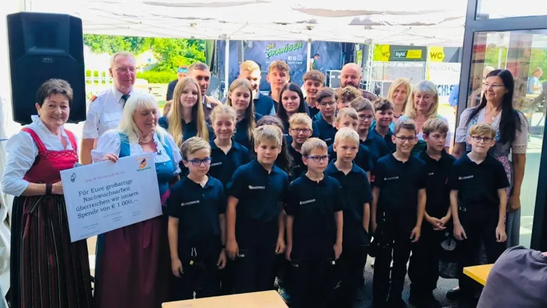 Eine Gruppe junger Schüler und Lehrer versammelt sich unter einem Zelt, lächelt und posiert für ein Foto. Eine Frau hält ein Stück Papier mit Text. Im Hintergrund ist ein großes blaues Banner mit Logos zu sehen.