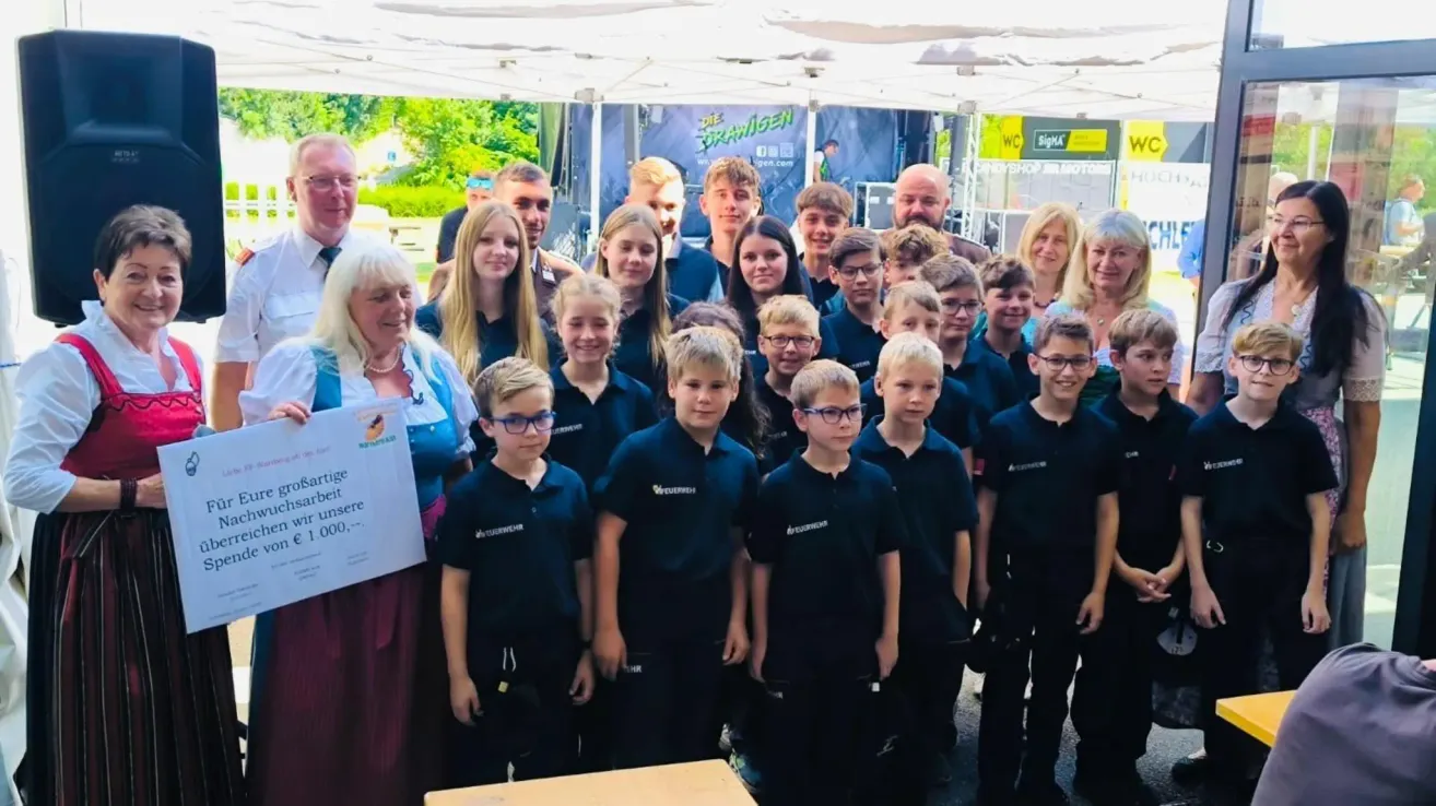 Eine Gruppe junger Schüler und Lehrer versammelt sich unter einem Zelt, lächelt und posiert für ein Foto. Eine Frau hält ein Stück Papier mit Text. Im Hintergrund ist ein großes blaues Banner mit Logos zu sehen.