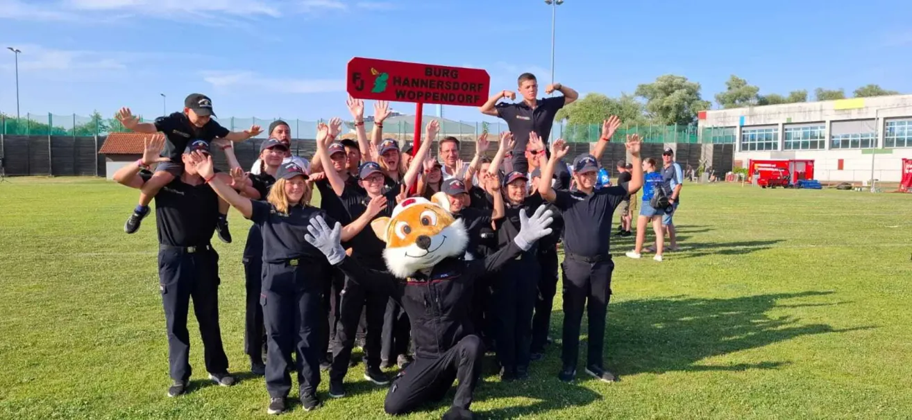 Eine Gruppe von Menschen in schwarzen Uniformen und Mützen steht in einer Reihe, einige mit erhobenen Händen. Eine Person in einer Fuchsmaske kniet vor ihnen, und ein rotes Schild steht für 'Burg Hannersdorf Woppendorf.' Ein Pfosten steht hinter ihnen.