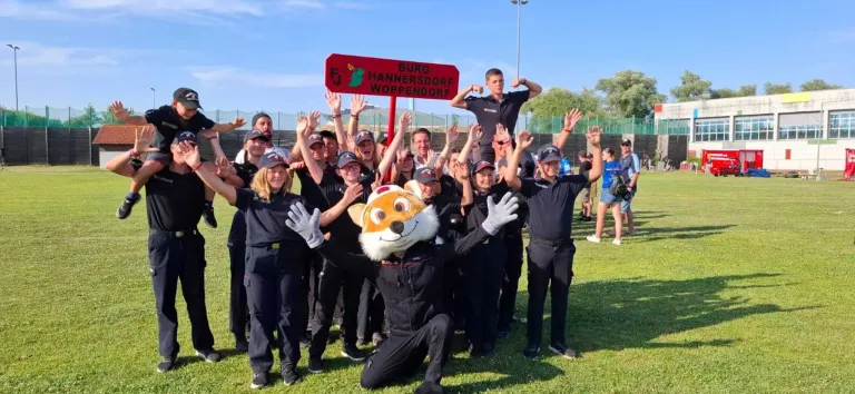 Eine Gruppe von Menschen in schwarzen Uniformen und Mützen steht in einer Reihe, einige mit erhobenen Händen. Eine Person in einer Fuchsmaske kniet vor ihnen, und ein rotes Schild steht für 'Burg Hannersdorf Woppendorf.' Ein Pfosten steht hinter ihnen.