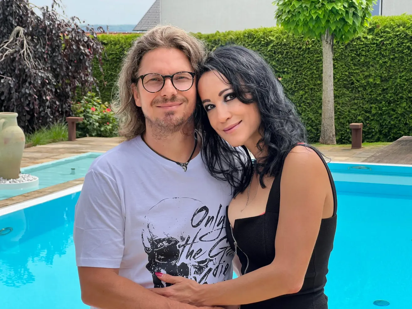 Ein Mann und eine Frau stehen neben einem Pool und lächeln für ein Foto. Der Mann trägt eine Brille und ein weißes T-Shirt mit Grafikdesign.