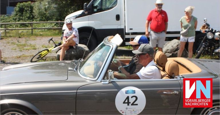 Ein altes Auto mit der Nummer 42 wird von einem Mann gefahren, während zwei andere neben ihm sitzen. In der Nähe ruht ein Radfahrer auf einem Felsen, und ein Mann in einem roten Hemd beobachtet. Ein weißer Lieferwagen ist dahinter geparkt.