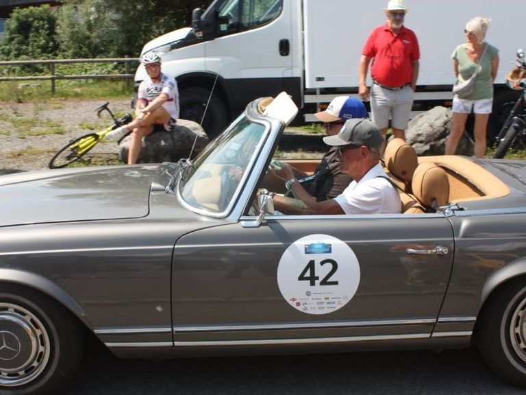 Ein grauer Cabriolet mit der Nummer 42 an der Tür. Zwei Männer sitzen im Auto, einer fährt. Hinter dem Auto sitzt ein Radfahrer auf einem Felsen. Ein Mann und eine Frau stehen in der Nähe, und ein weißer Lieferwagen ist dahinter geparkt.