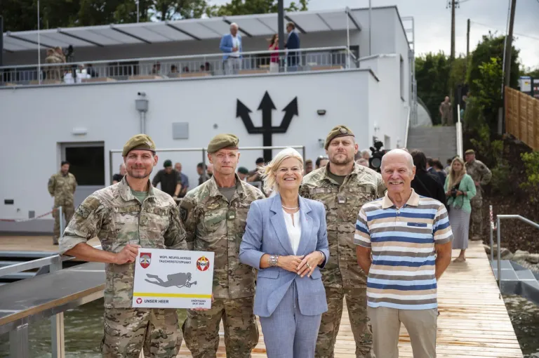 Fünf Personen, darunter zwei in Militäruniformen, stehen vor einem Gebäude mit einer Menge dahinter. Eine Frau in einem blauen Anzug hält ein Plakat, während zwei Männer auf beiden Seiten Kampfanzüge tragen.
