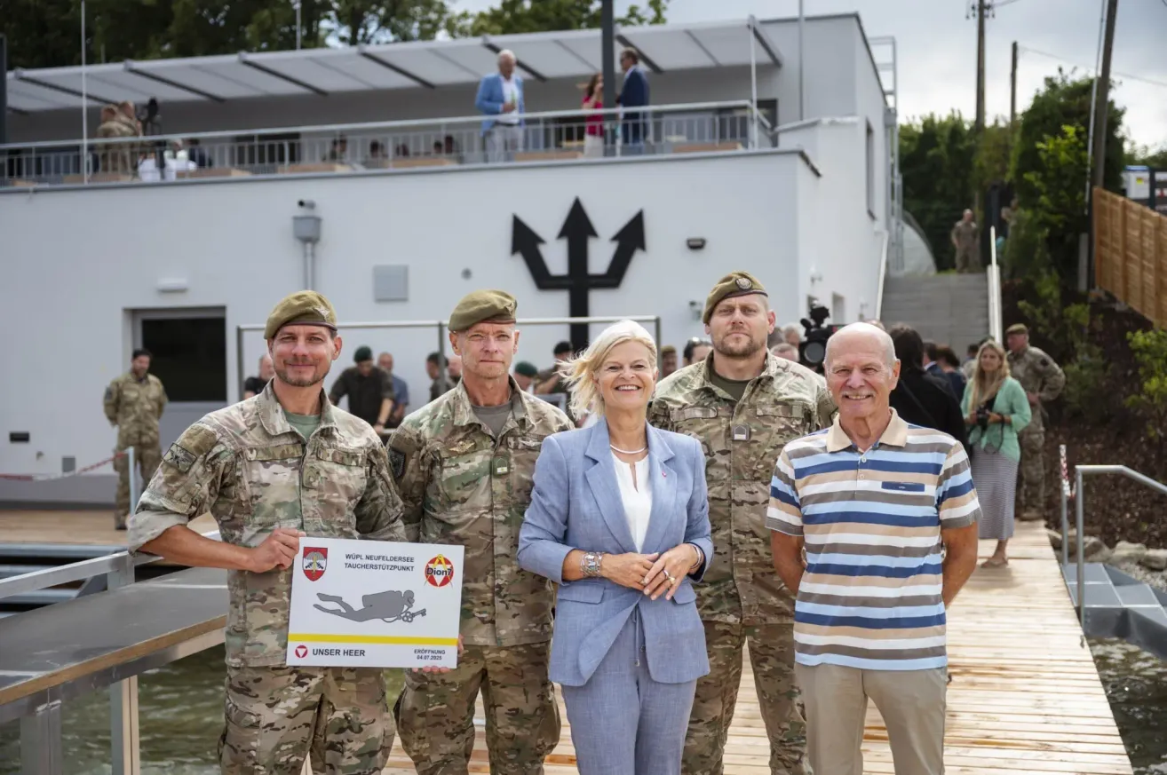 Fünf Personen, darunter zwei in Militäruniformen, stehen vor einem Gebäude mit einer Menge dahinter. Eine Frau in einem blauen Anzug hält ein Plakat, während zwei Männer auf beiden Seiten Kampfanzüge tragen.