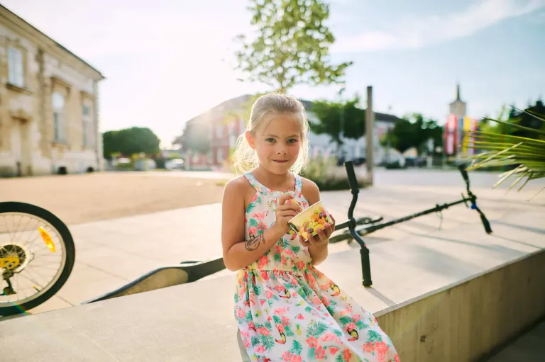 Ein junges Mädchen mit einem Tattoo auf dem Arm sitzt auf einer Betonkante und hält einen Becher mit Obst. Sie lächelt und scheint ihren Snack zu genießen. Hinter ihr sind ein Baum und Gebäude zu sehen.