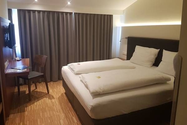 Ein Hotelzimmer mit einem Doppelbett, einem Schreibtisch, einem Stuhl und einem montierten Fernseher. Das Bett hat zwei Kissen und zwei Decken. Der Raum hat Holzboden.