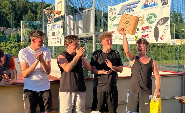 Eine Gruppe junger Männer steht bei einer Freiluft-Sportveranstaltung zusammen, einer hält eine Trophäe, jubelt und lächelt.