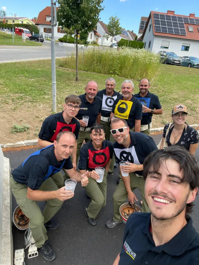 Eine Gruppe von Menschen in passenden Shirts posiert für ein Foto auf der Straße, hält Gläser in den Händen und lächelt. Hinter ihnen stehen Häuser mit Autos, die davor geparkt sind.