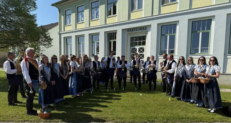 Eine Gruppe von Menschen in traditioneller Kleidung, einige mit Musikinstrumenten, steht vor einem Gebäude mit der Aufschrift 'Volksschule'.