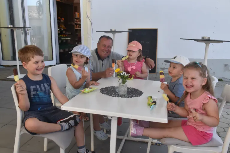 Eine Familie sitzt um einen Tisch draußen, mit einem Mann und vier Kindern, die lächeln. Jedes Kind hält eine Eisstange. Der Tisch hat eine Blumenvase und eine Tischdecke. Dahinter befindet sich ein Geschäft mit Regalen voller Artikel.