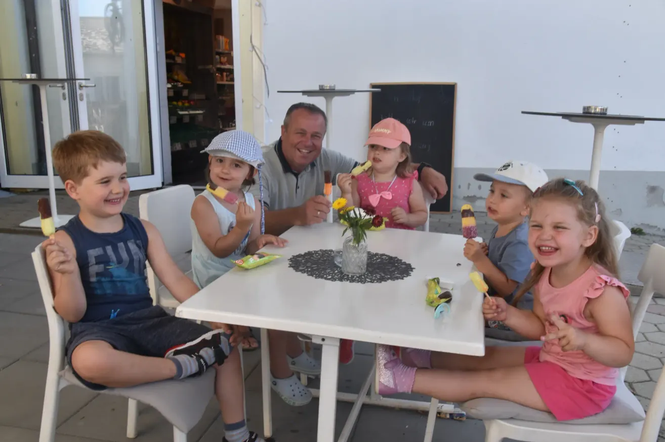 Eine Familie sitzt um einen Tisch draußen, mit einem Mann und vier Kindern, die lächeln. Jedes Kind hält eine Eisstange. Der Tisch hat eine Blumenvase und eine Tischdecke. Dahinter befindet sich ein Geschäft mit Regalen voller Artikel.