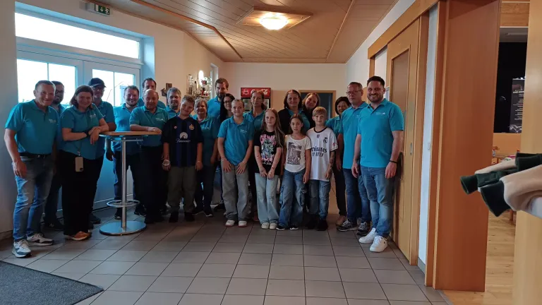 Eine Gruppe von Menschen, einschließlich Kindern, posiert für ein Foto in einem Raum mit gefliesten Böden und Holztüren. Einige tragen blaue Hemden, während andere Freizeitkleidung tragen.