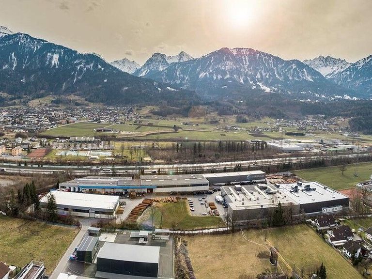 Luftaufnahme eines Industriekomplexes mit Gebäuden, Bergen und einer Stadt im Hintergrund. Das Gebiet ist von Grünflächen umgeben und durchquert von einer Eisenbahnlinie.