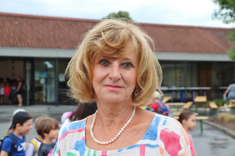 Eine Frau mit kurzen blonden Haaren trägt eine farbenfrohe Bluse und eine Perlenkette. Sie steht vor einem Gebäude mit Glasfenstern. Im Hintergrund spielen Kinder.