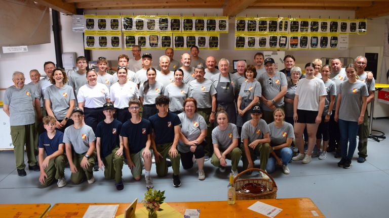 Eine Gruppe von Menschen in gleichen Uniformen posiert für ein Foto, einige stehen und andere knien. Sie befinden sich in einem Raum mit Holzboden und Postern an der Wand. Vor ihnen steht ein Korb und eine Flasche auf einem Tisch.