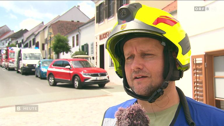 Ein Feuerwehrmann steht vor einem roten SUV, trägt einen gelben Helm und ein Mikrofon. Hinter ihm befinden sich Gebäude mit Fenstern und eine Straße.