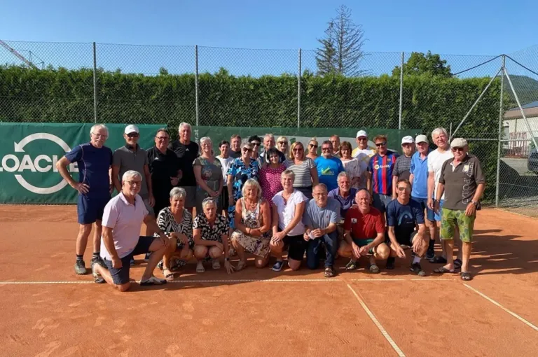 Eine Gruppe von Menschen in Freizeitkleidung posiert für ein Foto auf einem Tennisplatz mit einer weißen Linie. Dahinter befindet sich ein grüner Zaun mit Bäumen.