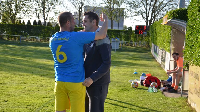 Zwei Männer auf einem Feld, einer in einem blauen Trikot mit der Nummer 6, der andere in einem schwarzen und grauen Jacke, stehen nah beieinander und schauen in eine Richtung.