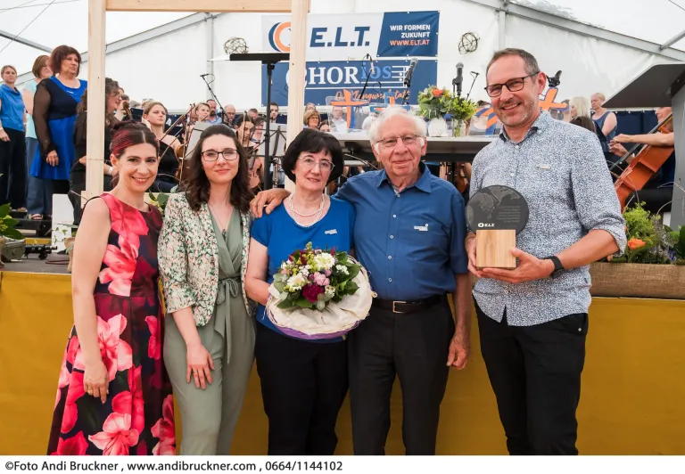 Vier Personen stehen lächelnd vor einem gelben Hintergrund und werden von einem Publikum umgeben. Hinter ihnen befindet sich eine Bühneneinrichtung. Ein Mann hält einen Preis und eine Frau eine Blumenstrauß.