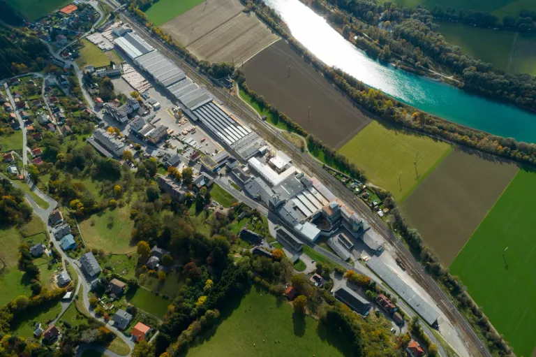 Ein Luftbild einer Fabrik am Flussufer mit Feldern und Häusern in der Umgebung. Gebäude, Autos und Grünflächen sind sichtbar.