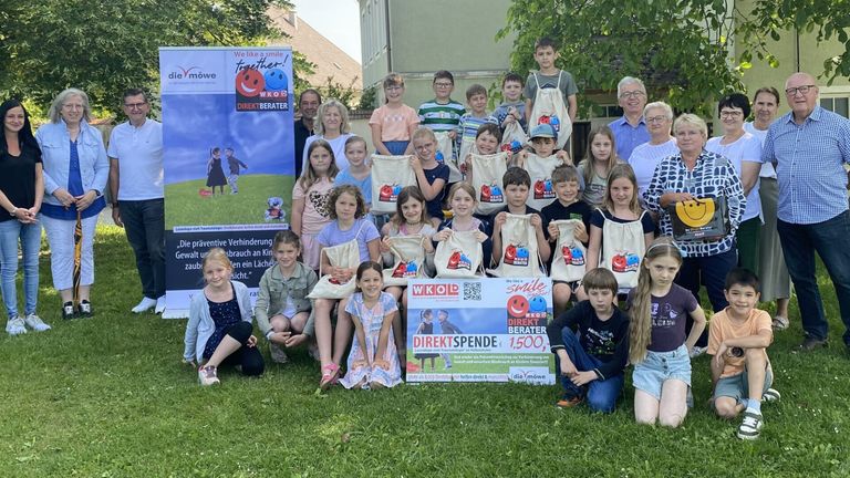 Eine Gruppe von Kindern und Erwachsenen lächelt für ein Foto im Freien. Sie halten Taschen mit dem Text 'We like a smile together' und 'DIREKT BERÄTER'. Dahinter befindet sich ein Plakat mit demselben Text. Einige von ihnen tragen Brillen und einige tragen Mützen.