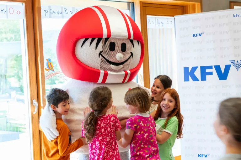 Eine Gruppe von Kindern steht um einen großen Maskottchen herum, das einen roten Helm und ein lächelndes Gesicht trägt, in einem Raum mit einem Schild im Hintergrund.