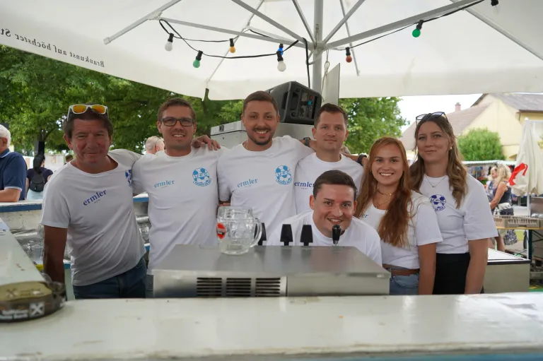 Eine Gruppe von Menschen in weißen T-Shirts posiert für ein Foto unter einem Zelt mit einem Schirm. Sie lächeln und stehen hinter einem Tresen mit einem Glaskrug.