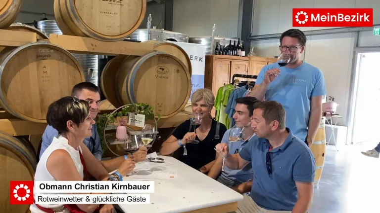 Eine Gruppe von Menschen sitzt in einem Weingut um einen Tisch herum und genießt Wein. Hinter ihnen sind Weinfässer und Flaschen zu sehen.
