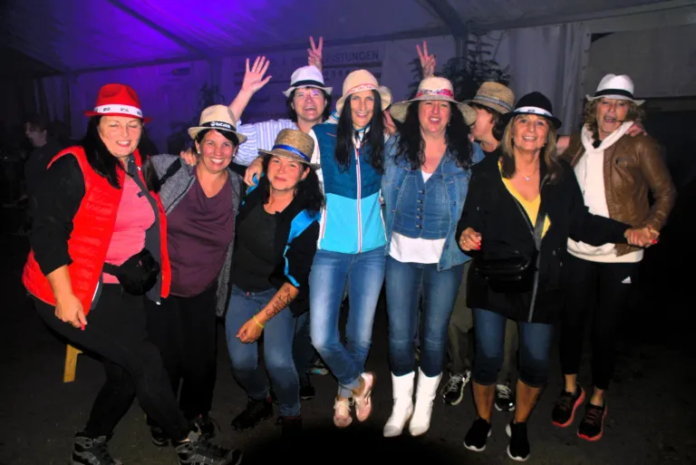 Eine Gruppe von Frauen in Hüten posiert für ein Foto unter einer violetten Beleuchtung. Einige tragen Jeans, andere Jacken. Sie stehen eng beieinander und lächeln.