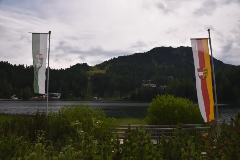Ein ruhiger Seeblick mit Bergen in der Ferne. Zwei Fahnen stehen im Vordergrund, eine mit einem Logo und die andere mit weißem Hintergrund.