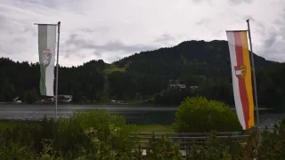 Ein ruhiger Seeblick mit Bergen in der Ferne. Zwei Fahnen stehen im Vordergrund, eine mit einem Logo und die andere mit weißem Hintergrund.