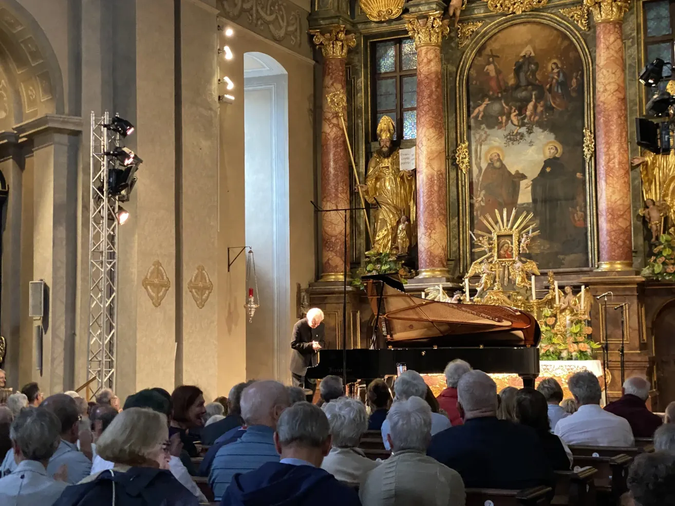 Ein älterer Mann spielt Klavier in einer Kirche, vor ihm sitzt ein Publikum. Hinter dem Klavier steht eine goldene Statue. Die Kirchenwände sind mit großen Gemälden verziert.