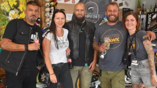 Fünf Personen posieren für ein Foto in einem Geschäft, mit Motorrädern im Hintergrund. Eine Frau in einem weißen T-Shirt hält eine schwarze Handtasche. Ein Mann in einer schwarzen Lederjacke steht neben ihr. Ein anderer Mann hält ein Glas. Die Wände des Geschäfts sind mit verschiedenen Aufklebern und einem großen Schild verziert.