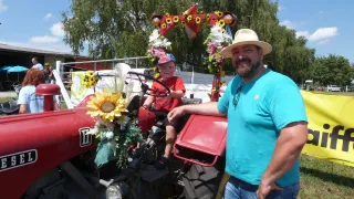 Ein Mann steht neben einem dekorierten Traktor, auf dem ein Kind im Fahrersitz sitzt, umgeben von Sonnenblumen und Blumen. Sie befinden sich auf einem Feld mit Bäumen und einem Gebäude im Hintergrund.