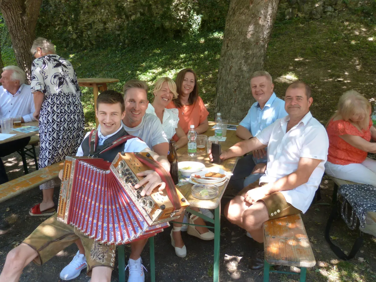 Eine Gruppe von Menschen sitzt lächelnd an einem Tisch im Freien. Ein Mann hält ein Akkordeon, während ein anderer eine Bierflasche hat. Teller mit Essen, Flaschen und ein Handy sind auf dem Tisch. Hinter ihnen steht eine Frau, und ein Baum bietet Schatten.